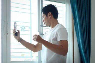 Asyalı bir adam, sabahları yatak odasının penceresinde durup badem sütü içerken selfie çekmek için cep telefonu kullanıyor. Akıllı telefonlu bir adam uyandıktan sonra sağlıklı bir içecek bardağı tutuyor..