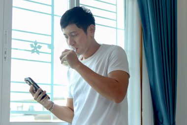 Asyalı adam badem sütü içerken akıllı telefon kullanıyor, sabahları yatak odasının penceresinde dikiliyor. Adam evde uyandıktan sonra sağlıklı içecek bardağını içerken cep telefonunu kontrol ediyor..