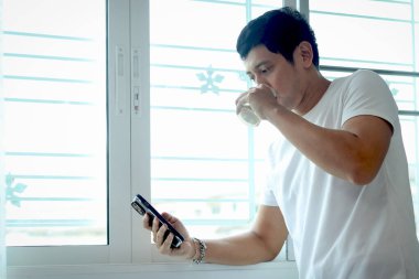 Asyalı adam badem sütü içerken akıllı telefon kullanıyor, sabahları yatak odasının penceresinde dikiliyor. Adam evde uyandıktan sonra sağlıklı içecek bardağını içerken cep telefonunu kontrol ediyor..