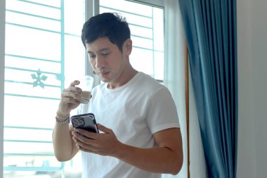 Asyalı adam badem sütü içerken akıllı telefon kullanıyor, sabahları yatak odasının penceresinde dikiliyor. Adam evde uyandıktan sonra sağlıklı içecek bardağını içerken cep telefonunu kontrol ediyor..
