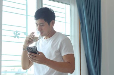 Asyalı adam badem sütü içerken akıllı telefon kullanıyor, sabahları yatak odasının penceresinde dikiliyor. Adam evde uyandıktan sonra sağlıklı içecek bardağını içerken cep telefonunu kontrol ediyor..