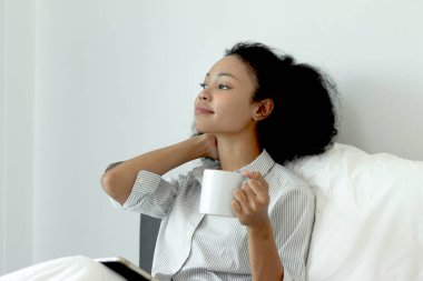 Kıvırcık saçlı güzel Afrikalı kadın sabah yatak odasında uyandıktan sonra tablet kullanırken kahve içiyor. Afrikalı kadın kahve fincanı tutuyor, rahat bir yatakta uzanırken tablet üzerinde çalışıyor..