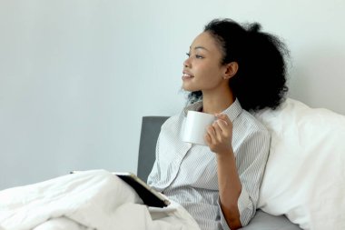 Kıvırcık saçlı güzel Afrikalı kadın sabah yatak odasında uyandıktan sonra tablet kullanırken kahve içiyor. Afrikalı kadın kahve fincanı tutuyor, rahat bir yatakta uzanırken tablet üzerinde çalışıyor..