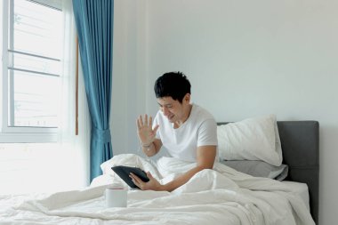 Asyalı adam sabah yatak odasında uyandıktan sonra yatakta tablet kullanırken el sallıyor. Tablet üzerinde çalışan bir adam, yatakta yatarken online toplantı yapıyor. Uykulu adam internette sörf yapıyor..