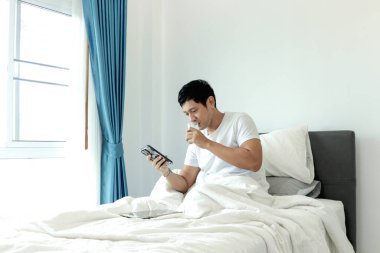 Asyalı bir adam sabah yatak odasında uyandıktan sonra yatakta badem sütü içerken akıllı telefon kullanıyor. Adam rahat bir yatakta uzanırken cep telefonunu kontrol ediyor. Uykulu adam internette sörf yapıyor.