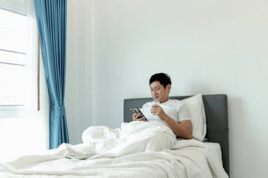 Asyalı bir adam sabah yatak odasında uyandıktan sonra yatakta badem sütü içerken akıllı telefon kullanıyor. Adam rahat bir yatakta uzanırken cep telefonunu kontrol ediyor. Uykulu adam internette sörf yapıyor.