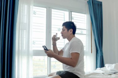 Asyalı adam badem sütü içerken akıllı telefon kullanıyor, yatak odasının penceresinin yanında oturuyor. Adam sabah evde uyandıktan sonra sağlıklı içecek bardağı içerken cep telefonunu kontrol ediyor.