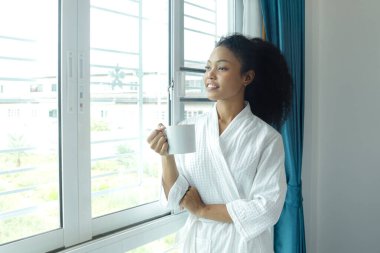 Beyaz bornozlu güzel kıvırcık saçlı kadın sabah kalktıktan sonra yatak odasında kahve fincanı tutuyordu. Mutlu Afrikalı kadın kahve içiyor, uyandıktan sonra kendini yenilenmiş hissediyor.