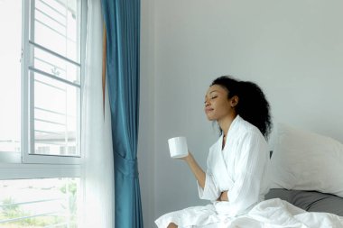 Beyaz bornozlu güzel kıvırcık saçlı kadın sabah kalktıktan sonra yatakta kahve fincanı tutarken. Mutlu Afrikalı kadın kahve içiyor, uyandıktan sonra kendini yenilenmiş hissediyor.