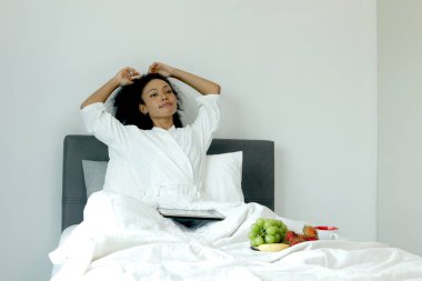 Beyaz bornozlu güzel kıvırcık saçlı kadın uyandıktan sonra tabletten gelen bilgileri kontrol ederken yatak odasında meyve kahvaltısı yapıyor. Mutlu Afrikalı kadın yatakta dijital cihaz kullanıyor