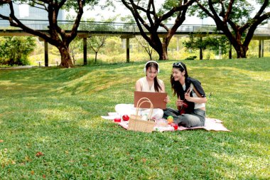 Parkta piknik yapan iki güzel Asyalı kadın. Yakın arkadaşlar ukulele oynar, açık havada yeşil bahçede piknik yaparken resim çizerler. İnsanlar doğal ortamlarda birlikte vakit geçirirler..