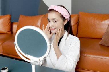 Bornozlu mutlu orta yaşlı Asyalı kadın aynaya bak, yüzüne kozmetik sür. Güzel, yetişkin bir kadın evde yüzüne cilt bakımı yapıyor. Güzellik Sağlığı ve Kozmetik.