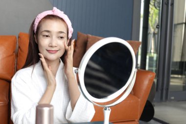 Bornozlu mutlu orta yaşlı Asyalı kadın aynaya bak, yüzüne kozmetik sür. Güzel, yetişkin bir kadın evde yüzüne cilt bakımı yapıyor. Güzellik Sağlığı ve Kozmetik.