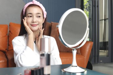 Bornozlu mutlu orta yaşlı Asyalı kadın aynaya bak, yüzüne kozmetik sür. Güzel, yetişkin bir kadın evde yüzüne cilt bakımı yapıyor. Güzellik Sağlığı ve Kozmetik.