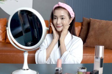 Bornozlu mutlu orta yaşlı Asyalı kadın aynaya bak, yüzüne kozmetik sür. Güzel, yetişkin bir kadın evde yüzüne cilt bakımı yapıyor. Güzellik Sağlığı ve Kozmetik.