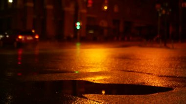Gece caddesindeki arabaların hareketlerinin arka planında bir su birikintisi. Asfalt seviyesinde hafif bir yağmur sırasında çekim yapmak.