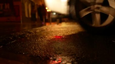Islak bir gece yolunda giden arabaların tekerlerini yakından çek. Arka planda odak dışı yayalar görünür.