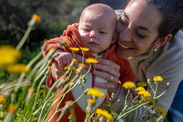 young smiling mother with her baby in the field touching the yellow flowers while going for a walk on a sunny day. baby discovering nature.