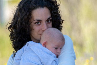 Kıvırcık saçlı genç anne yeni doğmuş bebeğini kollarında tutarken kameraya bakarak poz veriyor. aile ve yeni doğan bakım konsepti.