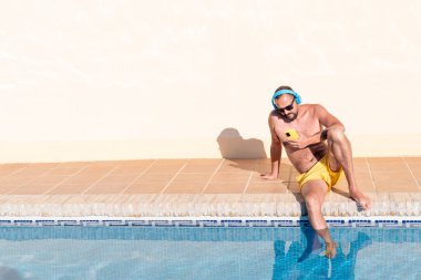Sakallı, sarı mayo ve güneş gözlüklü genç bir hippi mavi kulaklıklarıyla müzik dinlerken havuzda akıllı telefonuna bakıyor..