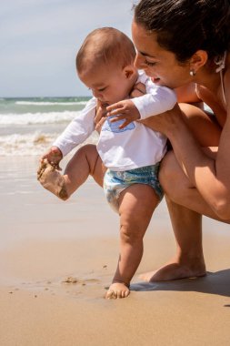 Genç bir anne ve bebeğinin sahilde ilk kez çekilmiş dikey fotoğrafı. Bebek ayağındaki kuma dokunuyor. Yazın çocuklarla plajda tatil kavramı