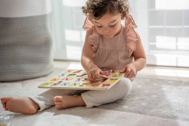 Odasındaki halıda oynayan kıvırcık saçlı, tahta alfabeyle oynayan Montessori kumaşlı küçük bir kızın yatay görüntüsü. çocukluk döneminde konsept geliştirme ve öğrenme.