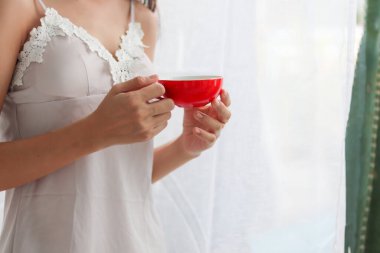 Seksi bir beyaz giyen genç bir kadın sabahları elinde kırmızı bir fincan kahveyle pencerenin kenarında durup güneşin doğuşunu seyrediyor..