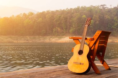 The wooden guitar was set up beside a chair on a wooden balcony in the morning over a reservoir with beautiful views of nature and bright sunshine in preparation for the upcoming party. clipart