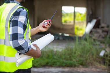 Perde arkasında, yansıtıcı yelek giyen ustabaşı inşaat alanının mühendislik ekibinin tasarım planlarına uyması ve sakinlerin güzellik ve güvenliği için incelenmesini sağlar..