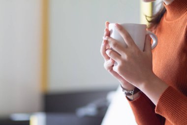 Kadın elinde sıcak kahve fincanı tutuyor ve yazlık evin dışındaki güzel manzaraya bakarken içiyor. Genç Asyalı kadın pencerenin kenarında sıcak kahve içiyor ve güzel manzaraya bakıyor.