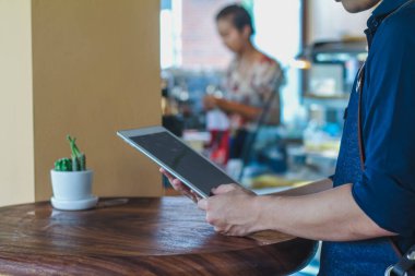 Küçük kahve dükkanının sahibi, hizmetlerinden faydalanmaya gelen müşterilerin siparişlerini beklemek için tableti elinde tutuyor. Önlüklü genç adam kahve dükkanında müşterilerden emir almayı bekliyor..