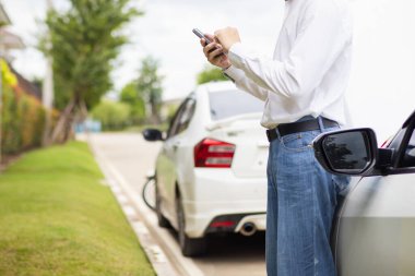 Delikanlı motor sorunu olan bir arabanın kaputunun önünde duruyor ve cep telefonuyla yerel bir araba servisini arıyor ve tamirciye gelip arabayı kontrol etmesini bildiriyor..