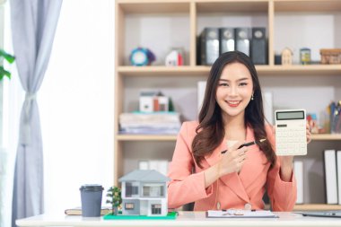 Asyalı emlakçı kadın ofiste oturmuş belgeleri hazırlıyor. İş kadını emlakçı çünkü artan nüfusla emlak işi büyümeye devam edecek..