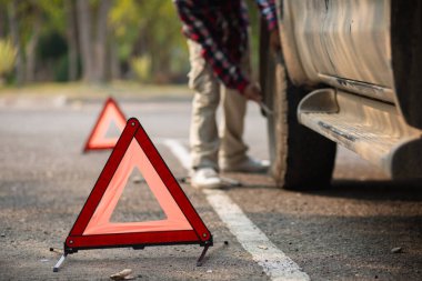 Bir adam, diğer sürücüleri yolun kenarına park etmiş arabalara dikkat etmeleri konusunda uyarmak için yansıtıcı bir üçgen kuruyor. Adamın biri patlak lastiği değiştirmek için yolun kenarına turuncu yansıtıcı bir üçgen kuruyor..