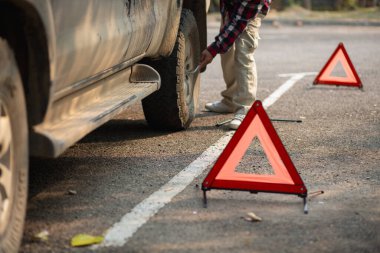 Bir adam, diğer sürücüleri yolun kenarına park etmiş arabalara dikkat etmeleri konusunda uyarmak için yansıtıcı bir üçgen kuruyor. Adamın biri patlak lastiği değiştirmek için yolun kenarına turuncu yansıtıcı bir üçgen kuruyor..