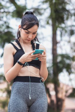 Sıkı spor giyimli Asyalı güzel koşucu tek başına ayakta duruyor ve moladan sonra müzik dinlemek için telefonunu açık tutuyor. Telefonunu tutan güzel kadınlar şarkıları seçer koşarken dinlerler