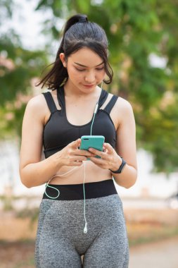 Sıkı spor giyimli Asyalı güzel koşucu tek başına ayakta duruyor ve moladan sonra müzik dinlemek için telefonunu açık tutuyor. Telefonunu tutan güzel kadınlar şarkıları seçer koşarken dinlerler