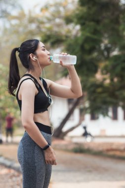 Plastik şişeden su içen güzel bir Asyalı koşucunun portresi. Çünkü koşmaktan yorgun hissediyor ve mola için su içmek istiyor. Güzel Asyalı kadın yorulduktan sonra su içiyor.