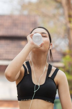 Plastik şişeden su içen güzel bir Asyalı koşucunun portresi. Çünkü koşmaktan yorgun hissediyor ve mola için su içmek istiyor. Güzel Asyalı kadın yorulduktan sonra su içiyor.