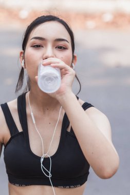 Plastik şişeden su içen güzel bir Asyalı koşucunun portresi. Çünkü koşmaktan yorgun hissediyor ve mola için su içmek istiyor. Güzel Asyalı kadın yorulduktan sonra su içiyor.
