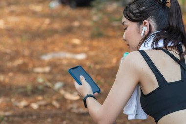 Sıkı spor giyimli Asyalı güzel koşucu tek başına ayakta duruyor ve moladan sonra müzik dinlemek için telefonunu açık tutuyor. Telefonunu tutan güzel kadınlar şarkıları seçer koşarken dinlerler