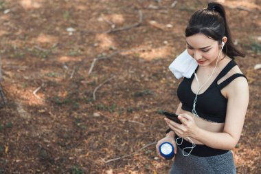 Sıkı spor giyimli Asyalı güzel koşucu tek başına ayakta duruyor ve moladan sonra müzik dinlemek için telefonunu açık tutuyor. Telefonunu tutan güzel kadınlar şarkıları seçer koşarken dinlerler