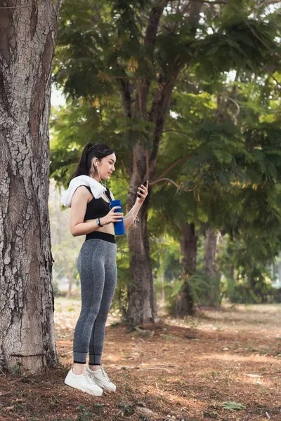 Sıkı spor giyimli Asyalı güzel koşucu tek başına ayakta duruyor ve moladan sonra müzik dinlemek için telefonunu açık tutuyor. Telefonunu tutan güzel kadınlar şarkıları seçer koşarken dinlerler