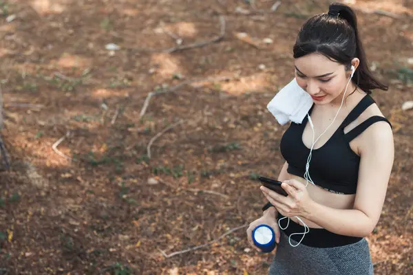 Sıkı spor giyimli Asyalı güzel koşucu tek başına ayakta duruyor ve moladan sonra müzik dinlemek için telefonunu açık tutuyor. Telefonunu tutan güzel kadınlar şarkıları seçer koşarken dinlerler