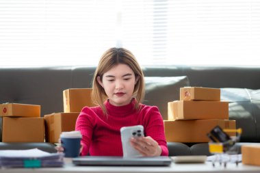 Kırmızı tişörtlü Asyalı iş kadını oturma odasının zemininde oturuyor siparişe göre müşterilere gönderilmeye hazır ürünleri paketlemek ve kutuları paketledikten sonra oturup dinlenmek için bekliyor..