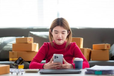 Kırmızı tişörtlü Asyalı iş kadını oturma odasının zemininde oturuyor siparişe göre müşterilere gönderilmeye hazır ürünleri paketlemek ve kutuları paketledikten sonra oturup dinlenmek için bekliyor..