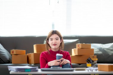 Kırmızı tişörtlü Asyalı iş kadını oturma odasının zemininde oturuyor siparişe göre müşterilere gönderilmeye hazır ürünleri paketlemek ve kutuları paketledikten sonra oturup dinlenmek için bekliyor..