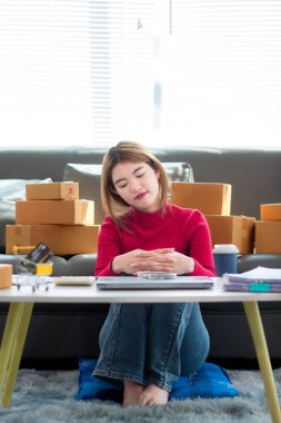 Kırmızı tişörtlü Asyalı iş kadını oturma odasının zemininde oturuyor siparişe göre müşterilere gönderilmeye hazır ürünleri paketlemek ve kutuları paketledikten sonra oturup dinlenmek için bekliyor..