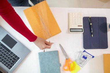 Kırmızı uzun kollu Asyalı bir iş kadını, çevrimiçi alışveriş uygulaması yoluyla yerleştirilen siparişlere göre ürünleri teslime hazır hale getirmek için uzun koli bandıyla paket paketleri hazırlıyor. Boşluğu kopyala