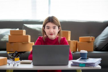 Kırmızı tişörtlü Asyalı iş kadını oturma odasının zemininde oturuyor siparişe göre müşterilere gönderilmeye hazır ürünleri paketlemek ve kutuları paketledikten sonra oturup dinlenmek için bekliyor..